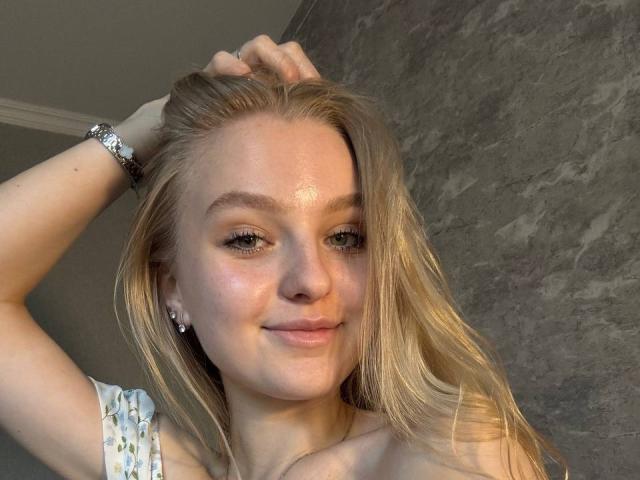 NievesMegee has blonde hair, fair skin, blue eyes, and a slight smile. She's holding her hair up, showing her tits and wearing a silver bracelet.