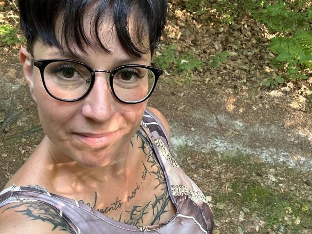TattoogirlDD-hot with short black hair, glasses, and tattoos on her chest, smiling at the camera, wearing a brown and white patterned top.