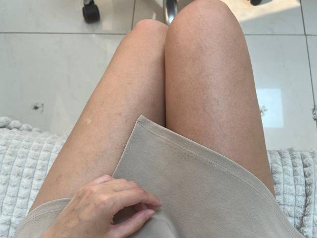 JuliannaMoor's knees are in the foreground, wearing a beige skirt, with a hand gripping the fabric. She's sitting on a white, textured blanket.