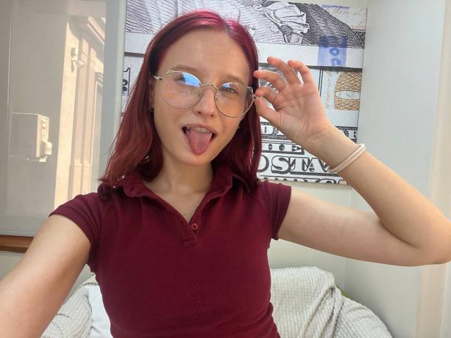 Julianna Moor, con cabello rojo, gafas y un polo color burdeos, sacando la lengua, sosteniendo sus gafas, una pulsera blanca y una cara linda, sentada en una cama..