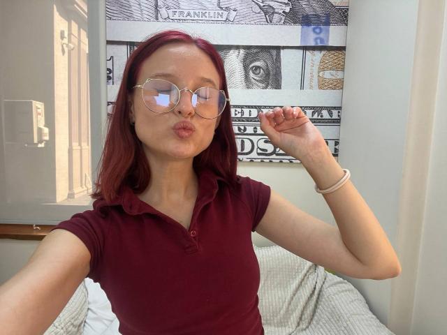 Julianna Moor, con cabello rojo, usando gafas y un polo color burdeos, frunciendo los labios, con la mano derecha levantada, un brazalete blanco en la muñeca, sentada en una cama, durante el día..