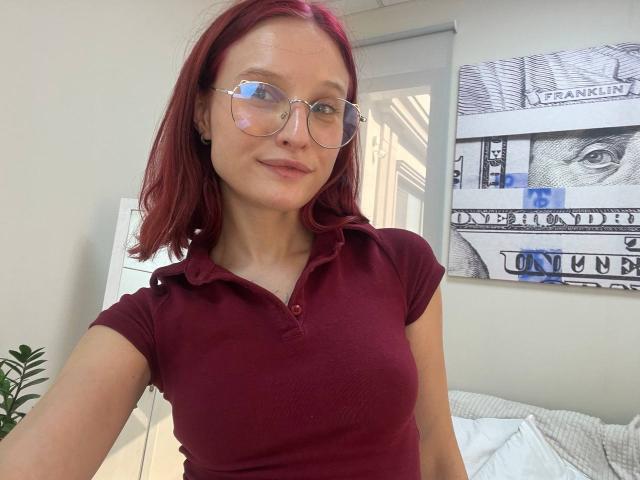 JuliannaMoor with red hair, glasses, and a maroon polo, showing her small tits in a selfie. She's smiling slightly, standing in a bright room with a plant.