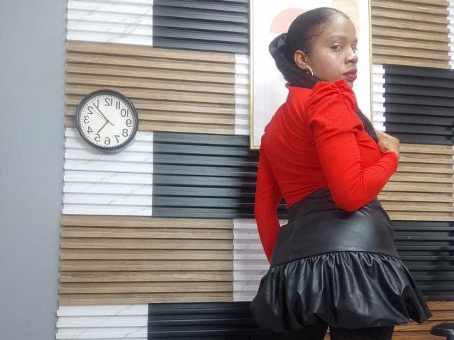 SarahiPusey has black skin, a red long-sleeve top, black leather skirt with a flared bottom, and her back is to the camera, looking over her shoulder. Clock on wall.