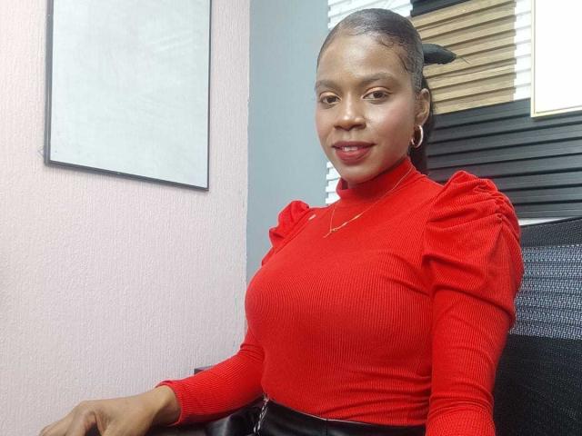 SarahiPusey with dark skin, small tits, and a red ribbed long-sleeve top, wearing black pants, hoop earrings, and a gold necklace, smiling at the camera.