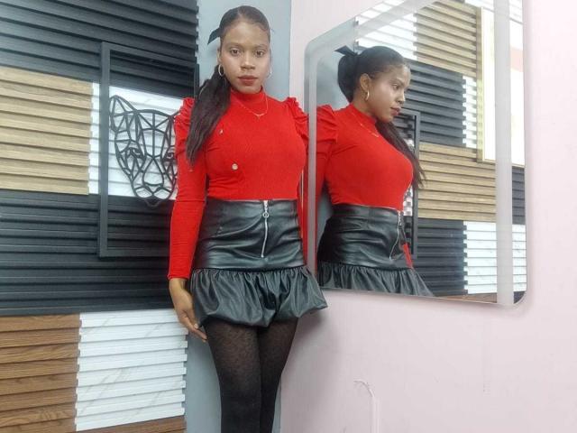 SarahiPusey with long black hair in a ponytail, wearing a red turtleneck, black leather skirt with ruffle, and black patterned tights, standing in front of a mirror.