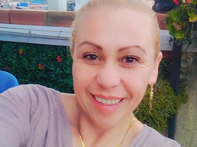 BiancaVelvet with blonde hair, blue eyes, and a big smile, wearing a purple shirt and gold earrings, showing her cute nose and shiny teeth, looking happy.