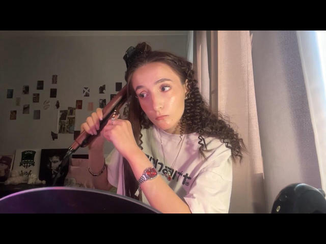 BrunildaSchweppe with curly brown hair, white shirt, and wrist bracelet, holding a brush in her right hand, looking to the side, in a dimly lit room.
