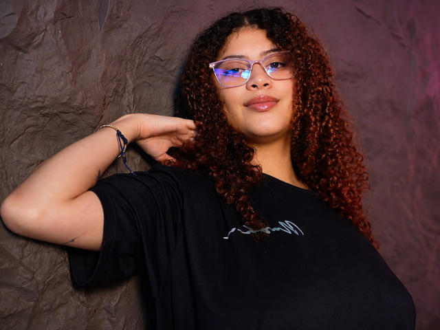 HoneyHipsss with curly brown hair, glasses, and a black t-shirt, leaning against a wrinkled brown wall, smiling with her arm raised, showing a bracelet. Boobs visible, small tattoo.