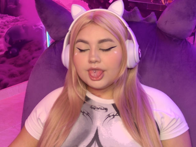 MarlaBriard with long blonde hair, white cat-ear headphones, eyes closed, tongue out, white t-shirt, and a lip piercing, sitting in a pink-lit room. Her tits are small.