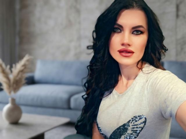 MilaLoona a de longs cheveux noirs ondulés, des yeux bleus et du rouge à lèvres. Elle porte une chemise grise avec un motif d'oiseau et est assise devant une table avec un vase en arrière-plan..