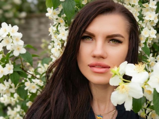 MilaLoona has green eyes, pink lips, and long brown hair, surrounded by white flowers. She's wearing a black top and a blue necklace. Her ass is not visible.