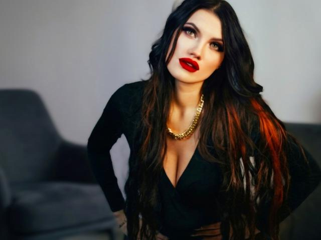 MilaLoona, avec ses longs cheveux noirs, son rouge à lèvres rouge, sa chaîne en or et son décolleté en V noir, mettant en valeur sa poitrine généreuse et son allure sexy et confiante. Elle se tient debout, les mains sur les hanches..
