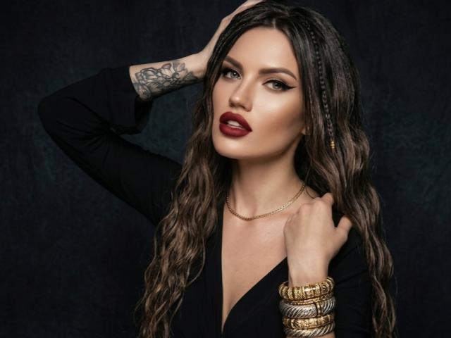 MilaLoona mit welligem, braunem Haar, rotem Lippenstift, schwarzem Oberteil, goldener Armbanduhr und tätowiertem Arm, Hand im Haar, blickt in die Kamera, dunkler Hintergrund, sexy, selbstbewusst, attraktiv..