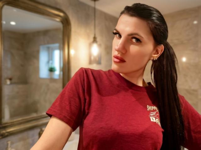 MilaLoona with dark ponytail, red shirt, and earrings, looking at camera with red lipstick, showing her tits slightly. Warm bathroom lighting, gold-framed mirror in background.