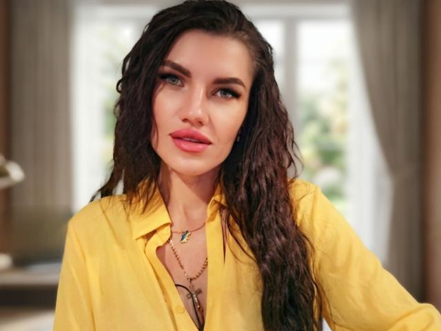MilaLoona, met nat, donkerbruin haar, een gele blouse en een ketting met een kleine blauwe hanger, heeft roze lippen en een subtiele, sexy uitstraling..