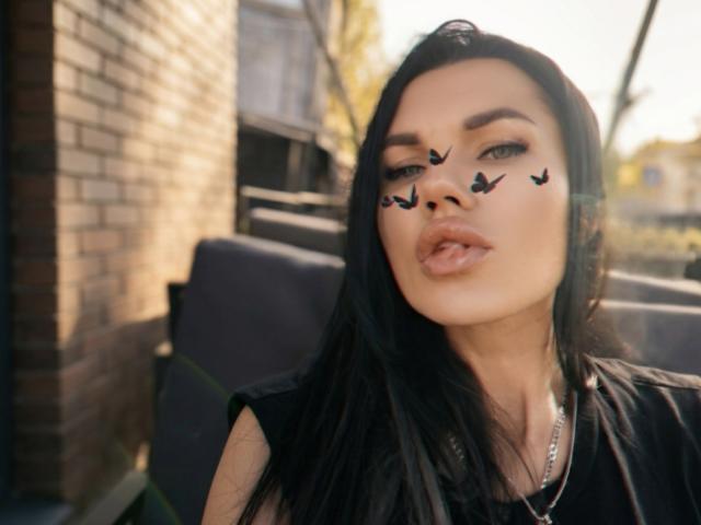 MilaLoona mit schwarzen Haaren, Make-up mit Fledermausflügeln auf der Nase, voller Lippen, trägt ein schwarzes Oberteil, eine silberne Halskette, und im Hintergrund ist Sonnenschein zu sehen..