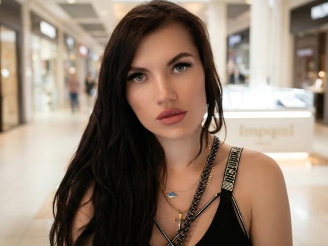 MilaLoona hat langes, schwarzes Haar, blaue Augen und roten Lippenstift. Sie trägt ein schwarzes Oberteil mit einer Kettenkette und einem kleinen Kreuzanhänger. Ihre Brüste sind klein..