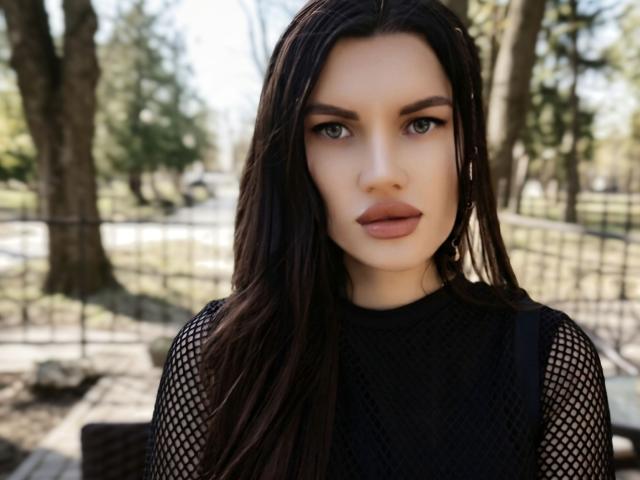 MilaLoona with long dark brown hair, wearing a black mesh top, showing her tits through the net, staring directly at the camera with a neutral expression.