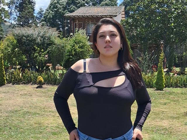 PaulaAnd has dark brown hair, medium tits, and a black cut-out top showing a black bra strap, standing in a grassy yard, wearing blue jeans.