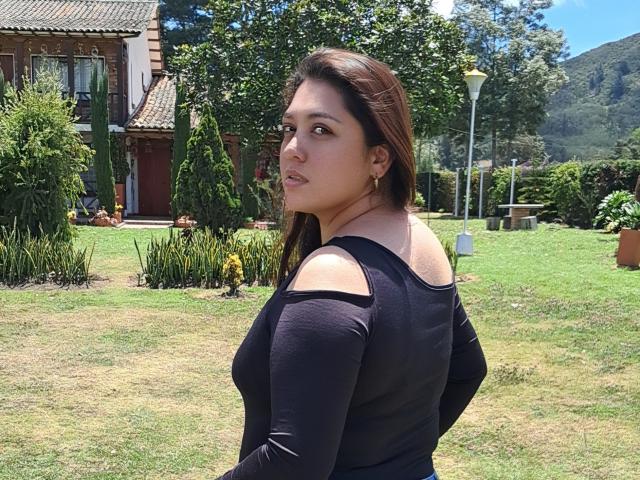 PaulaAnd has brown hair, wearing a black, long-sleeve top with cut-out shoulders, standing on grass, looking back over her shoulder, with a serious expression.