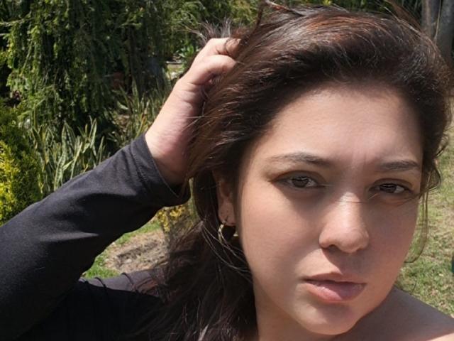 PaulaAnd has brown hair, tan skin, and is looking into the camera with a neutral expression, wearing a black long-sleeve shirt, hand in hair, outside in sunlight.