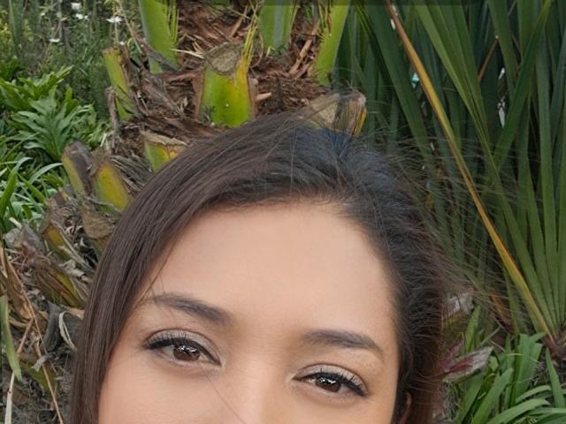 PaulaAnd's face with brown eyes, long eyelashes, and brown hair, partially hidden by palm leaves and green plants, in a natural outdoor setting.