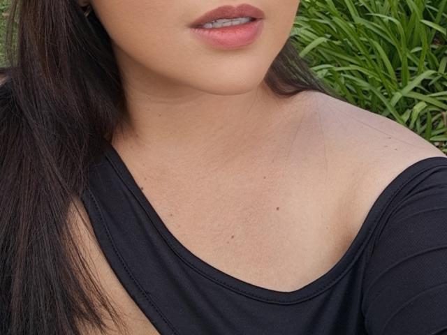 PaulaAnd's face and chest, showing her pouty pink lips and black off-shoulder top, with her brown hair and light skin, a few small moles visible on her skin.