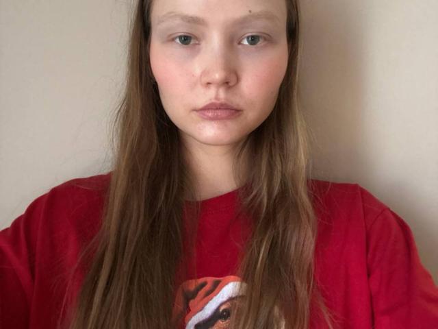 Eula Yanek hat braunes, glattes Haar, helle Haut und grüne Augen. Sie trägt ein rotes Hemd mit einem Teilmuster und blickt direkt in die Kamera, wobei sie einen neutralen Gesichtsausdruck hat..