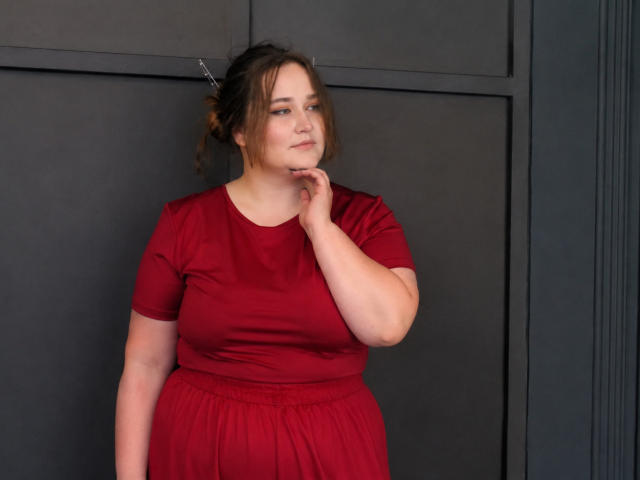 DaniellaGraceZZ, con la pelle chiara, i capelli castani raccolti in un disordinato chignon, un vestito rosso, una mano che tocca il mento, che guarda di lato, in piedi di fronte a una parete grigio scuro..