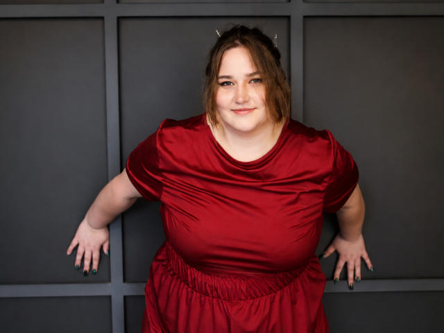 DaniellaGraceZZ with chubby tits, wearing a shiny red dress, leaning against a dark wall, smiling slightly, hands on either side, black nails, brown hair, subtle makeup.