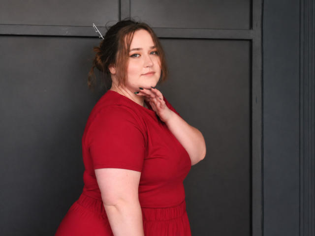 DaniellaGraceZZ with pale skin, brown hair in a messy bun, wearing a red dress, standing sideways, hand near her neck, smiling softly, showing her curvy, medium-ass and tits.