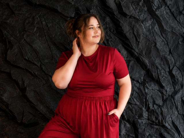 DaniellaGraceZZ mit wohlgeformten, mittelgroßen Brüsten und Gesäß, trägt ein rotes Kleid, eine Hand in der Tasche, vor einem dunklen, rauen Fels, lächelt leicht, braunes Haar zu einem unordentlichen Dutt gebunden..
