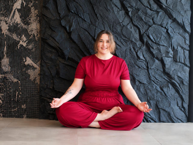 DaniellaGraceZZ, chubby with big tits, sits cross-legged in red clothes, smiling, hands in meditation pose, tattoo on left arm, against dark textured wall, bare feet, blonde hair.