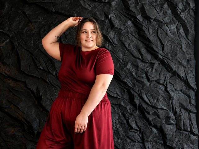 DaniellaGraceZZ has a plus-size body with big tits and a curvy ass, wearing a red dress, standing against a dark textured wall, smiling with one hand up.