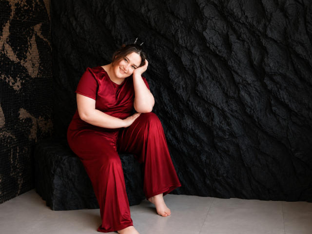 DaniellaGraceZZ with pale skin, brown hair, and a curvy ass, sits barefoot on a black bench wearing a red satin dress, smiling at the camera.