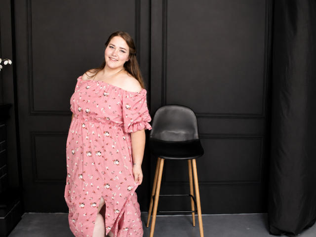 DaniellaGraceZZ, carina con un seno di media grandezza, indossa un abito floreale rosa con una spalla scoperta, in piedi accanto a una sedia nera con gambe in legno, sorridendo alla fotocamera..
