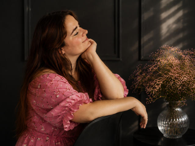 DaniellaGraceZZ with long brown hair, pink floral dress, hand on chin, pensive expression, sitting by black chair, vase with pink flowers on right. Soft light, relaxed mood.