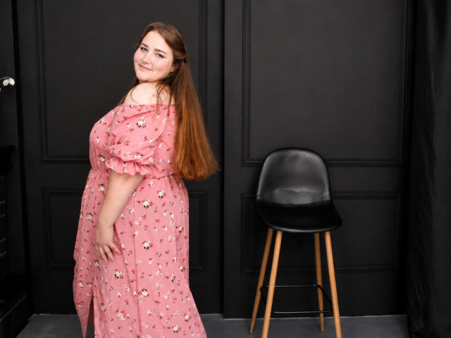 DaniellaGraceZZ con lunghi capelli castani, un abito floreale rosa, che mette in mostra il suo seno prosperoso, in piedi accanto a una sedia nera, sorridente, che guarda oltre la spalla..