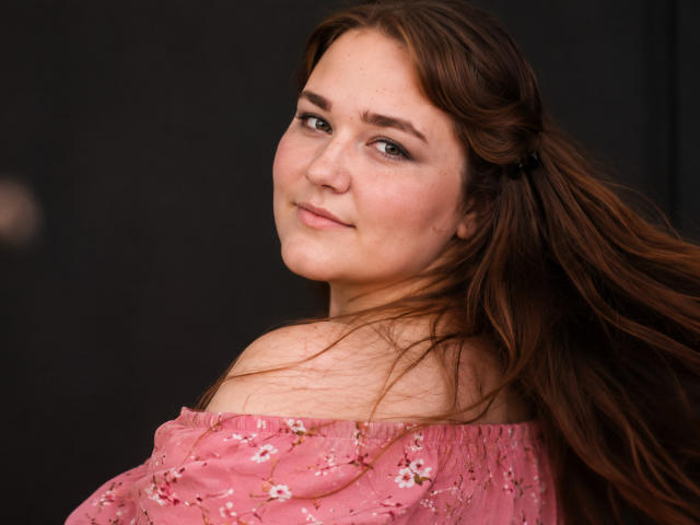 DaniellaGraceZZ with long brown hair, off-shoulder pink floral top, subtle smile, and light makeup, looking over shoulder, slight wind in hair, soft skin, pretty, cute.