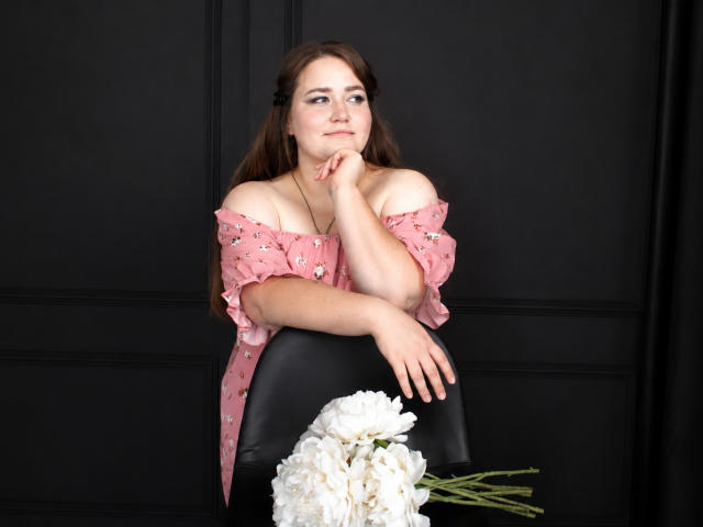 DaniellaGraceZZ with brown hair, pink off-shoulder floral top, resting chin on hand, black chair, white flowers on table, black wall, slight smile, relaxed pose.