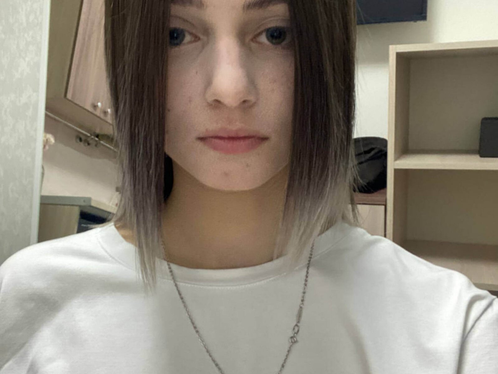 Miyoko Dolence hat braunes, glattes Haar mit grauen Spitzen, blaue Augen und Sommersprossen. Sie trägt ein weißes Hemd mit einer silbernen Halskette und blickt direkt in die Kamera..