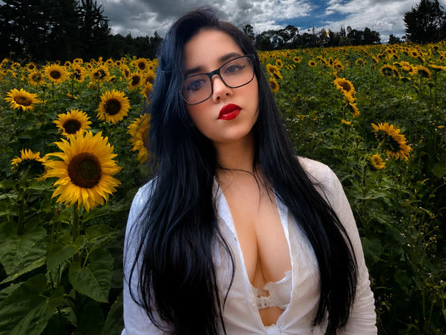 Kkatalina with long black hair, glasses, red lipstick, white open shirt showing her tits, standing in a sunflower field under a cloudy sky.