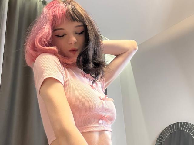 ChangCaddle with pink and black hair, wearing a pink crop top, has her arm raised, showing her small tits, and has her eyes closed in a soft, relaxed pose.