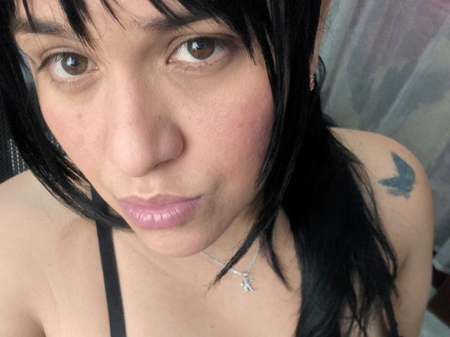 Viviana Thompson tem cabelo preto, olhos castanhos, lábios rosados, uma tatuagem preta no ombro direito e veste uma blusa preta com alças, com um colar de cruz prateado..