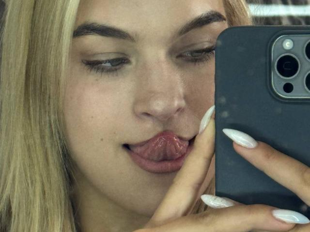AlinaLemon's face, blonde hair, winking eyes, tongue out, holding a phone with white nails, her lips slightly parted, and her nose centered in the shot.