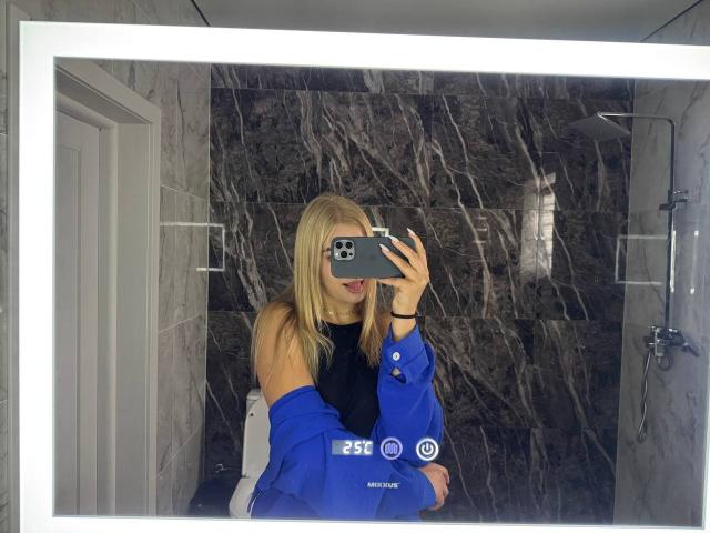 AlinaLemon with blonde hair, blue off-shoulder top, black sleeve, holding phone in mirror selfie, grey marble wall, white door, and shower in background, tongue slightly out.