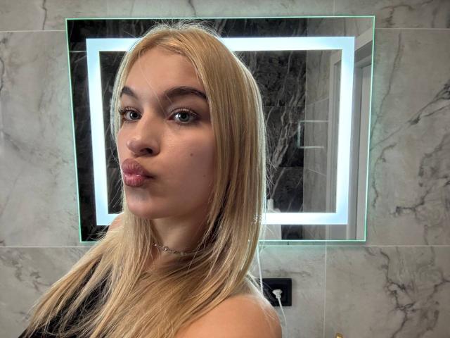Blond-haired AlinaLemon with pouty lips, wearing a necklace, in front of a lit mirror. Her green eyes and subtle makeup are visible. Her tits are not in the frame.