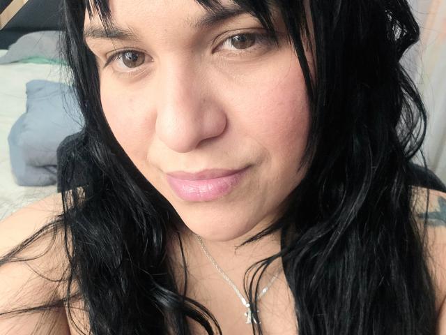 VivianaThompson has black, wavy hair, light skin, brown eyes, and pink lips. She's wearing a silver necklace with a cross, looking at the camera with a slight smile.