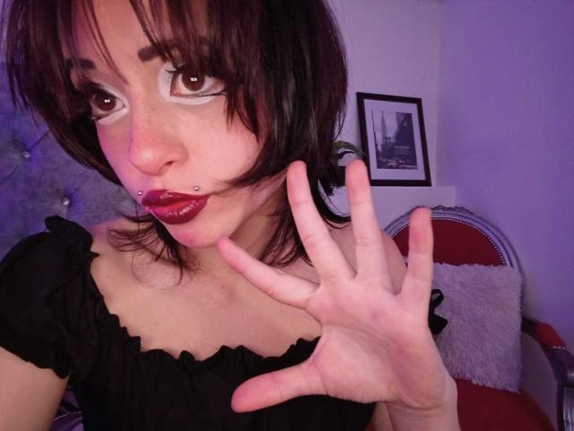 ViolettClay has dark brown eyes, white makeup around eyes, black shirt, red lipstick, nose piercing, and black hair. She's winking with her right hand up.