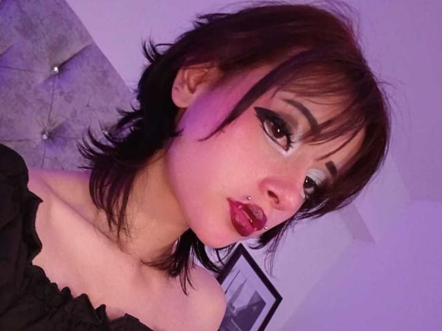 ViolettClay with dark brown hair, black eyeliner, pierced nose, red lipstick, and black off-shoulder top, showing her bare shoulders and small, perky tits.